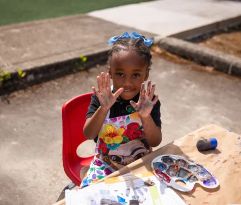 young child holding up their hands covered in paint at a table playing with paints