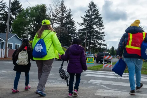 Two adults in brightly colored safety gear accompany two children across a crosswalk.