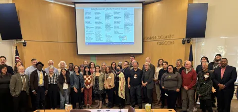a large group of various genders, races, ages, and clothing colors gathered at the front of the Multnomah County Board Room. A screen with about 100 names of honored volunteers.