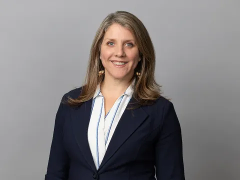 Smiling woman with long hair wearing a navy blazer and a white shirt against a gray background. She conveys a professional and approachable demeanor.