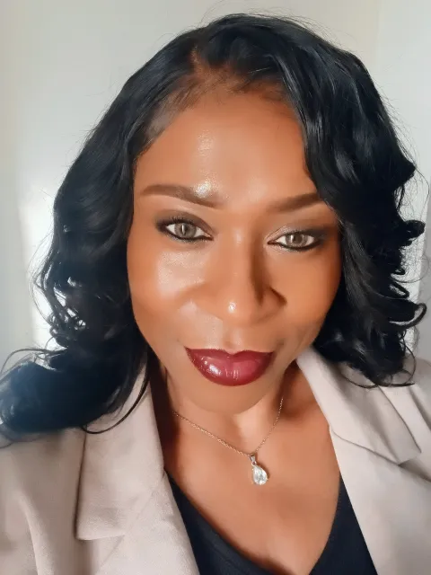 A woman with long, curly black hair smiles confidently at the camera. She's wearing a light blazer, a pendant necklace, and elegant dark lipstick, conveying a poised and professional tone.
