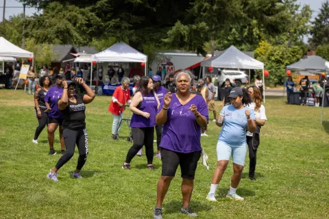 Un grupo de mujeres negras bailando en línea en un campo en una feria comunitaria con carpas al fondo.