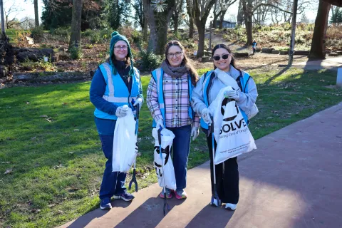 Three people holding SOLVE bags against a background of green grass and trees.