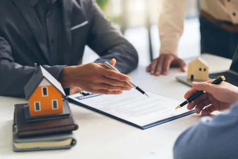 Three people discuss a real estate contract at a table with model houses. Professional setting, collaborative tone, and focus on documents.