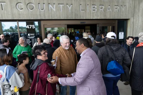 Multnomah County Commissioner Jones-Dixon, wearing a light purple suit jacket, smiling and shaking hands with community members in a crowd outside the entrance of the East County Library during its grand opening.