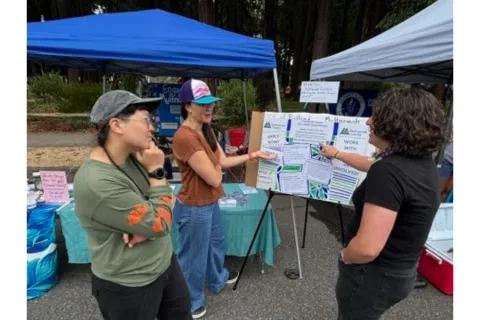 Three adults stand outdoors around a poster board with information about the Charter Review Process