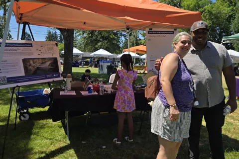 Two people stand smiling in front of an information table at an outdoor event.