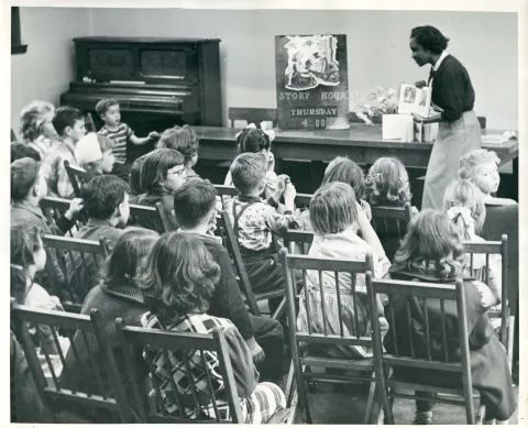 Hora del cuento en la sucursal de Arleta. La bibliotecaria Lois Tolbert Sayles está de pie leyendo un libro a un grupo de niños sentados en sillas de madera. Lois fue la primera bibliotecaria afroamericana en trabajar para la Biblioteca del Condado de Multnomah. Un cartel en una mesa de madera dice: «HORA DEL CUENTO, JUEVES, 4:00». Un piano vertical se encuentra en la esquina trasera izquierda.